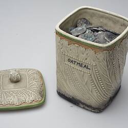 Fire affected cream ceramic container and lid that contains melted coins. Lid off.