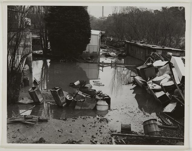 Flood damaged outdoor area with much debris and some remaining water. Trees nearby and chinmey in distant back