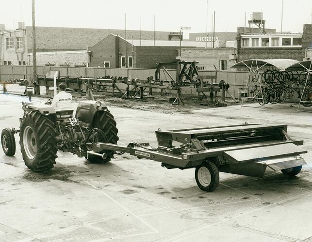 Man driving a tractor towing a windrower in factory grounds.