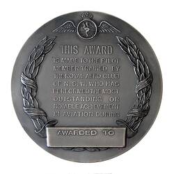 Dull silver round medal with wreath framing text.