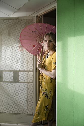 Person in yellow dress holding parasol stands in doorway.