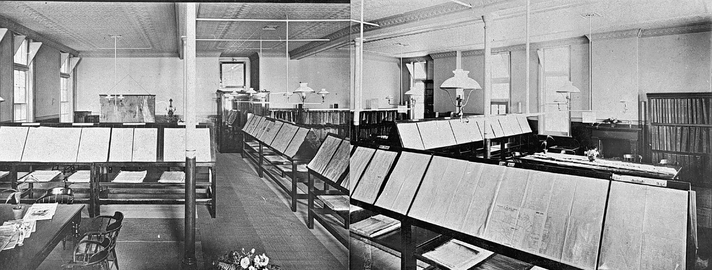 A newspaper reading room with slanted racks holding open papers A newspaper reading room with rows of slanted wooden racks holding open newspapers under hanging lamps