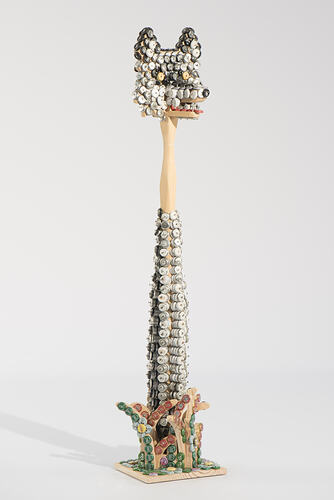 Tall thin instrument covered with bottle caps. Has bottle top head of wolf.