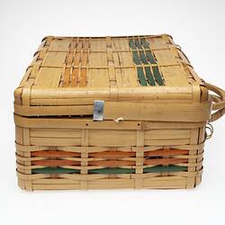 Cane basket, hinge lid. Orange and green stripe detail. Two handles at front. Profile.