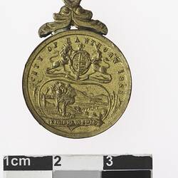 Round gold medal with coat of arms above trees and field, with text.