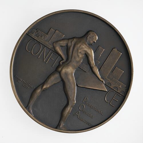 Round medal with nude male holding drawing board. City scene behind with ship and bridge.