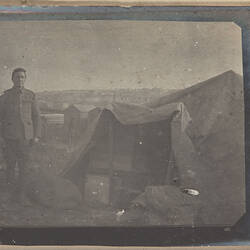 Soldier standing next to a tent, camp visible in background.