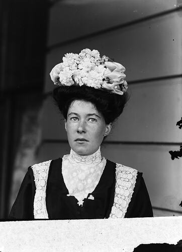 Woman posing in black dress with white lace collar. She wears a decorative white hat.