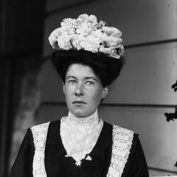 Woman posing in black dress with white lace collar. She wears a decorative white hat.