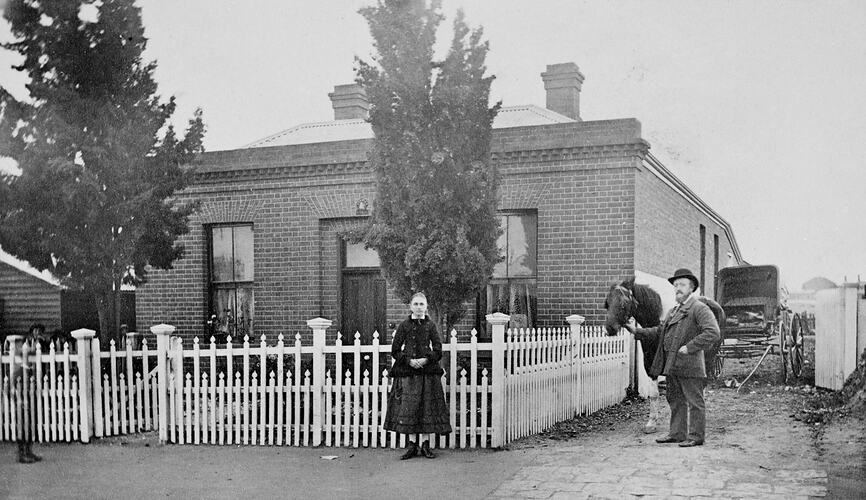 Man and girl in front of brick house with picket fence. He has a horse. Carriage in background.