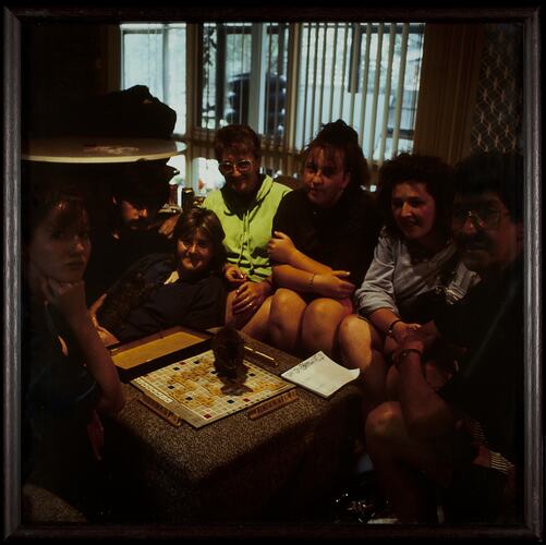 Group sitting in dim room around table playing Scrabble board game.