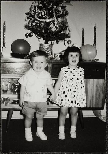 Small boy and girl holding hands in front of a Christmas tree.