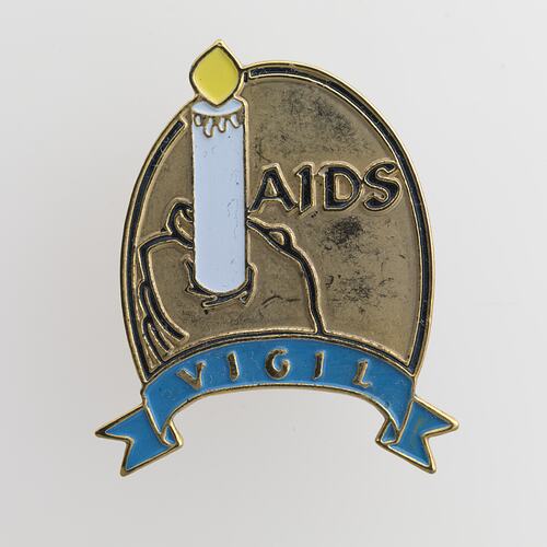 Gold coloured badge with white candle being held but hand. Blue scroll beneath. Raised text.