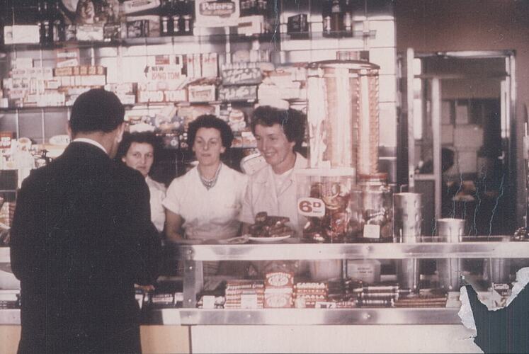 Three women at counter serving man.