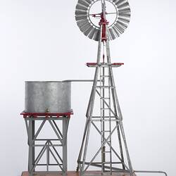 Model water pumping windmill next to water tank on a stand. Vanes facing rear.