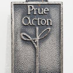Cast metal badge, raised text above a stylised letter 'T' logo.