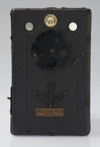 Black box-shaped camera with round metal plate and badges.