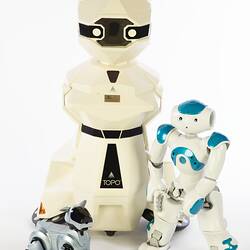 Cream plastic robot with angular human-like shape, ie. head, shoulders, waist and legs. Siver robot dog at left, smaller blue white root at right.