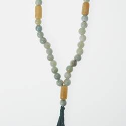 Beaded loop with pale blue, grey, yellow and white beads. Black tassel at end. At base.