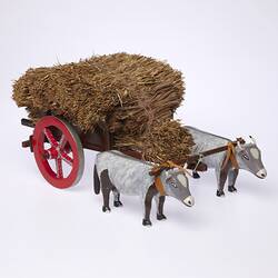 Model of two grey bullocks pulling a hay wagon with haystack and red wheels. Front three quarter view.