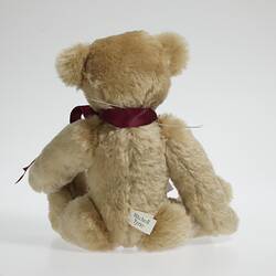 Back of seated light brown plush bear with red ribbon around neck.