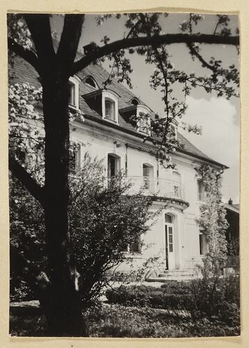 White double-storey house with balcony and attic windows. Tree in foreground.