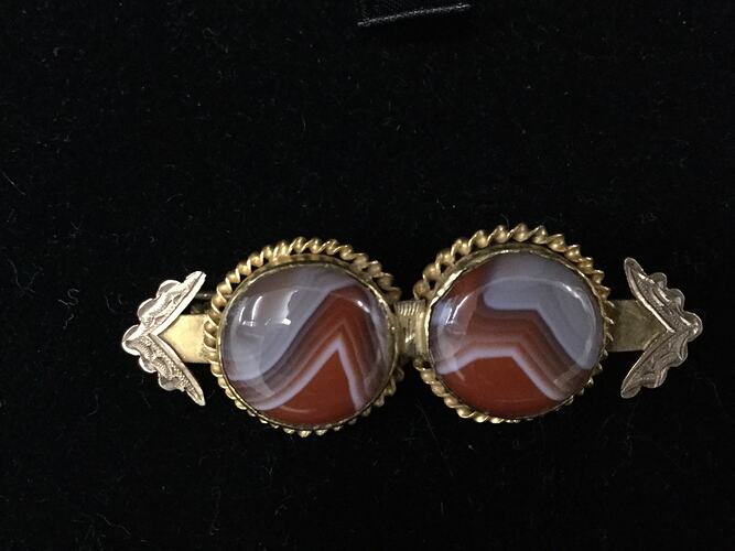Two side-by-side stone discs with multi-coloured layered patterning set on a gold bar brooch with jagged points at each end.
