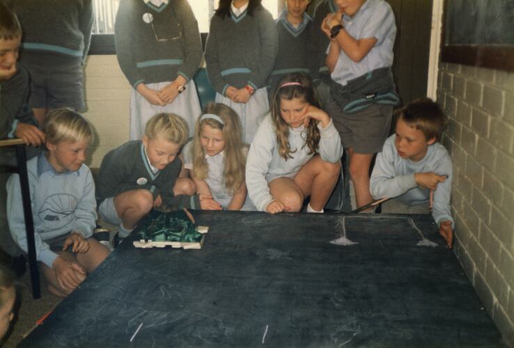 A group of children watching a Lego turtle moving around a table.