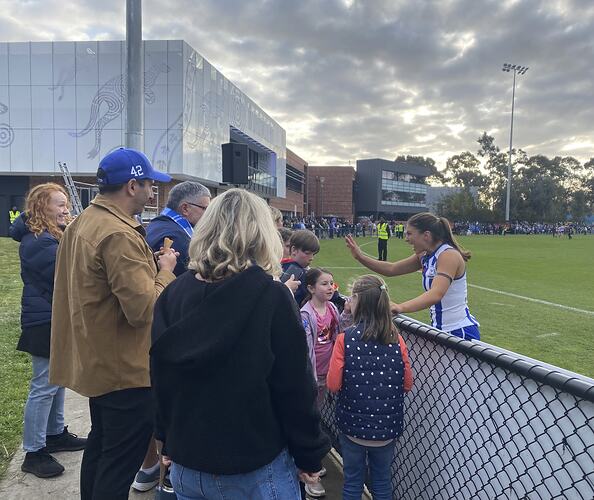 Spectators seen from behind football oval fence looking at/interacting with female footballer on oval.