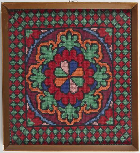 Framed square cloth embroidered with central 8-petalled floral motif in blue, green, yellow and light pink. Red, green and yellow patterns form border.