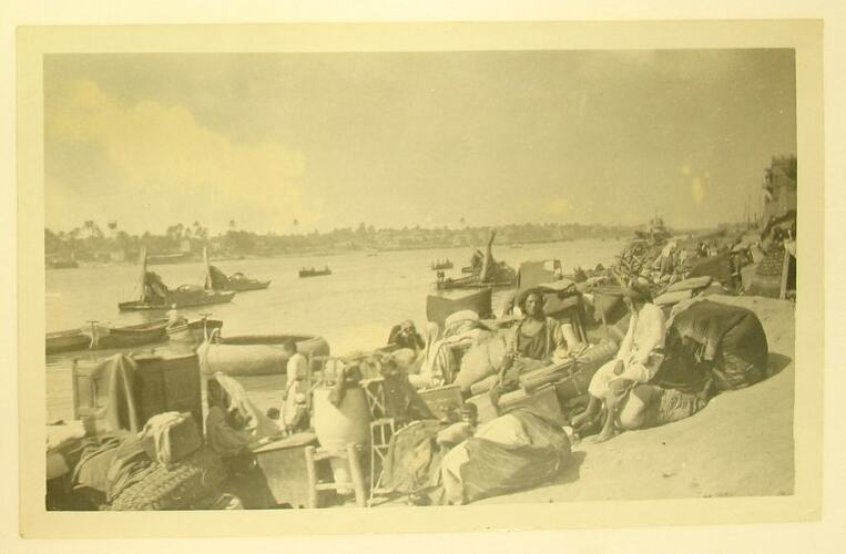 Men seated beside river surrounded by bags and furniture.