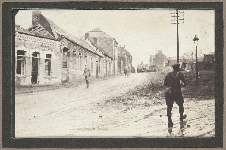 Street with bomb damaged building on the right side, three servicemen on the dirt road.