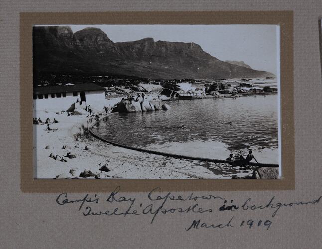 Bay with sun bathers and rocky outcrop, mountains in background.