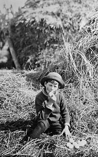 A boy, seated in a haystack, sucking on an egg.