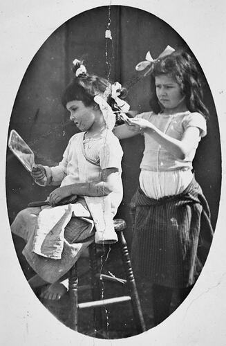 Standing girl 'doing' hair of another seated girl who holds a mirror.
