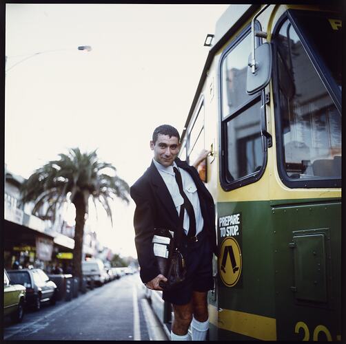 Man in uniform with tram ticket satchel hangs from side of green and yellow tram.