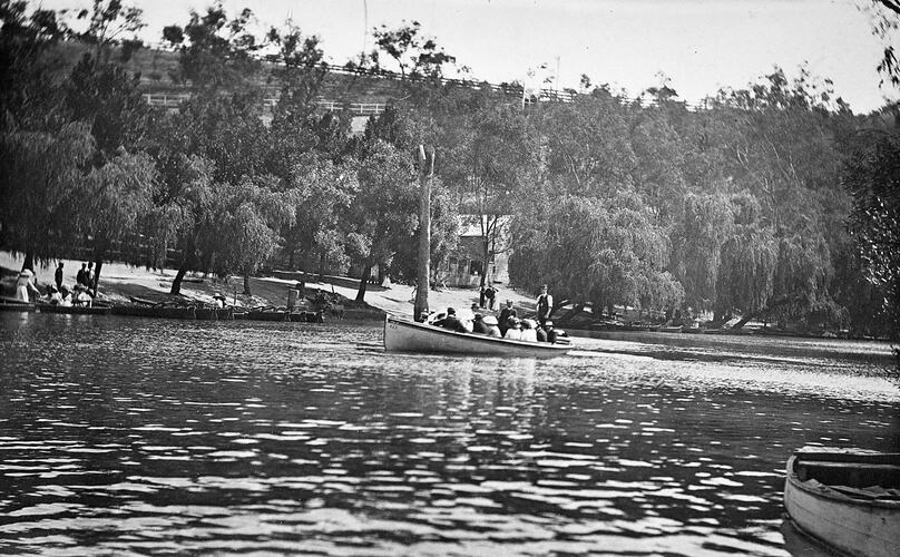 Boat on river with seated passengers. One man stands. Several boats on riverbank. Boat shed amongst trees in b