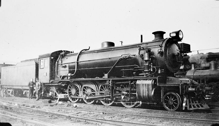 Two men, probably staff, stand beside a locomotive, which is shown in side view.