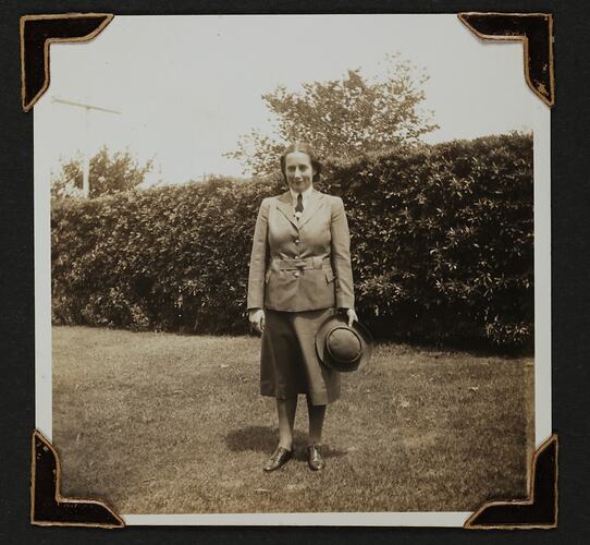 Woman in uniform standing in a yard with tree hedge behind.