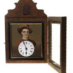 Wooden cuckoo clock with woman's portrait in gold frame. Clock face below her face. Glass door open.