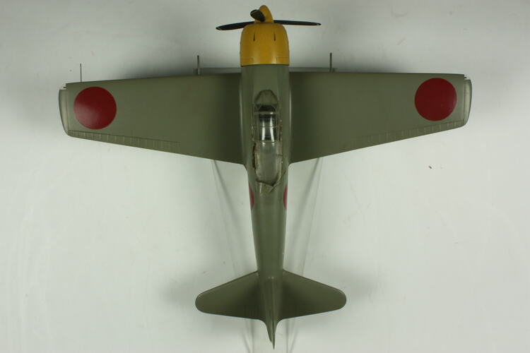 Green grey plane model with red spot on each wing and fusleage. Yellow nose, black propeller. Top view.