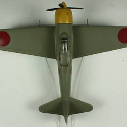 Green grey plane model with red spot on each wing and fusleage. Yellow nose, black propeller. Top view.