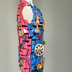 Right side of dress made of horizontal strips of pink, yellow, blue plastic. Adorned with plastic cables, doll