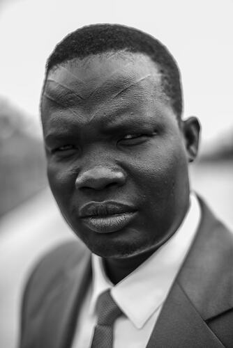 Man wearing dark suit jacket, tie and white shirt. He has V-shape markings on his forehead.