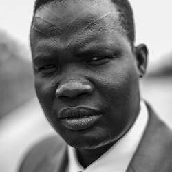 Man wearing dark suit jacket, tie and white shirt. He has V-shape markings on his forehead.