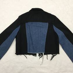 Back of short two-toned jacket made from blue and black denim.It has a collar, two pockets, long sleeves.