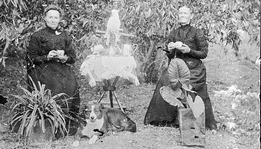 Two women holding teacups seated at a table in a garden. Cockatoo on perch behind table and dog below it.