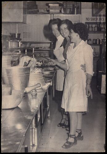 Three woman smiling whilst working in a kitchen. There are many dishes.