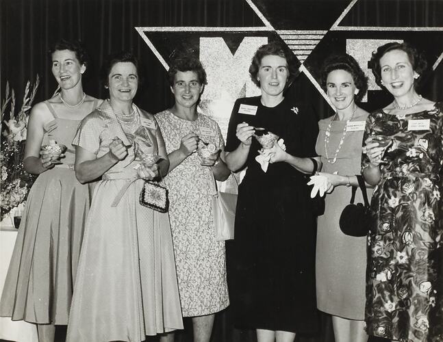 Six women in formal dress stand in a group at a Massey Harris Ferguson's dealers event.