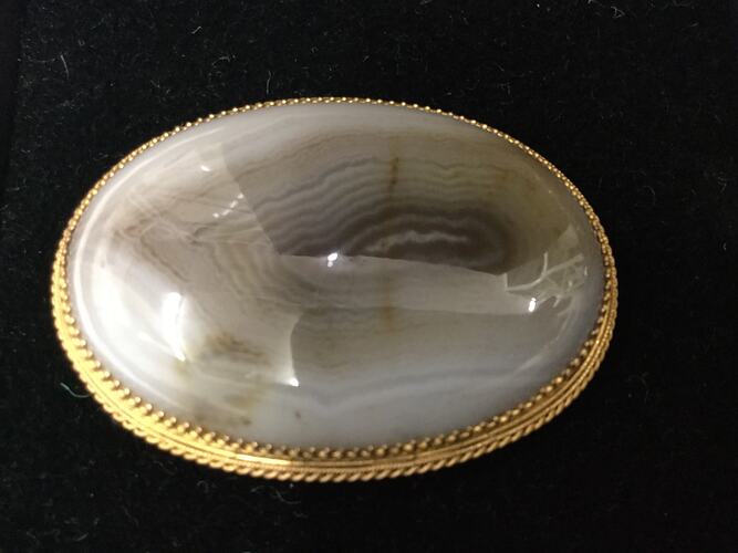 Polished oval stone brooch featuring creams and browns in a patterned gold setting.
